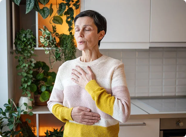 A woman stands with her eyes closed, practicing mindful breathing with her hands on her chest and stomach.