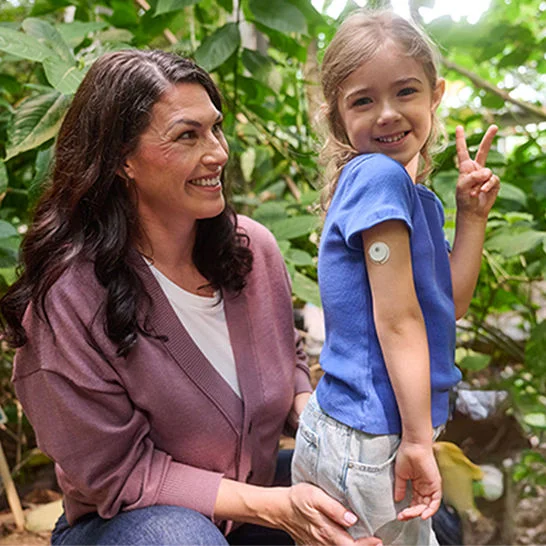 Child wearing a blue shirt shows a peace sign while displaying a glucose monitor on an upper arm, seated beside an adult in a lush garden.