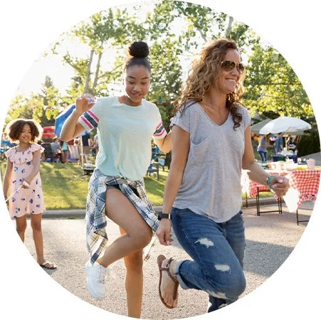 Three people skipping together on a sunlit street at an outdoor community event with tables and trees nearby.