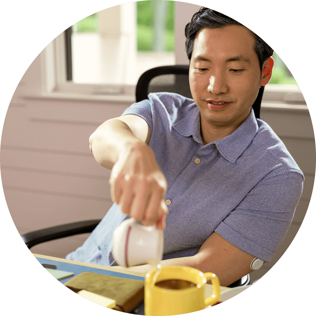 A man pours cream into his coffee at a desk, with a FreeStyle Libre sensor on his upper arm.