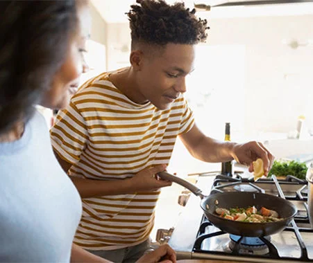 Person adding ingredients to a skillet on a stovetop while another person watches in a bright kitchen.