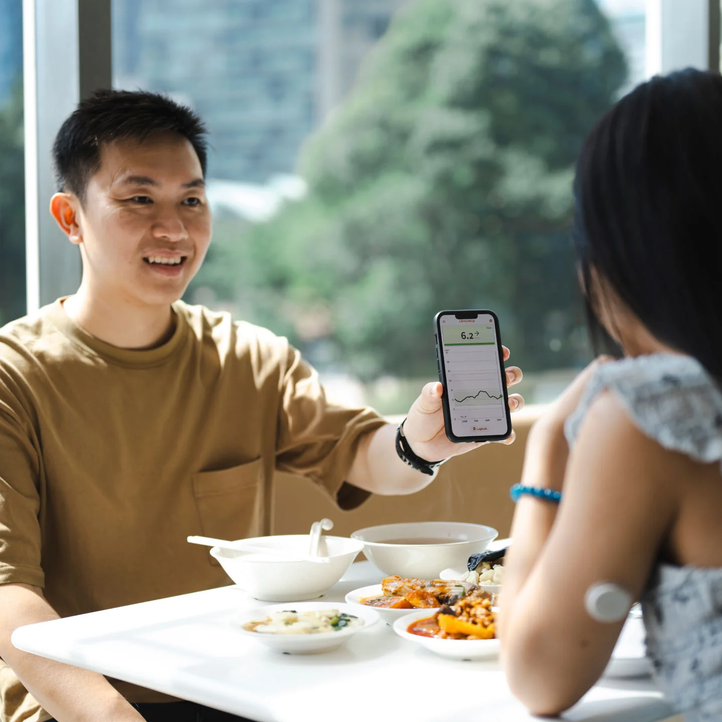 Young man shows an older woman wearing a FreeStyle Libre sensor on her upper arm something on his smartphone while they sit together on a couch.