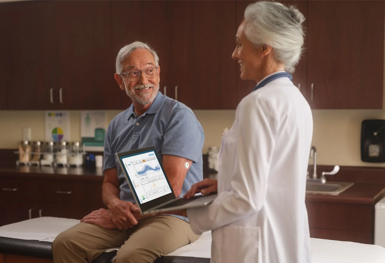 Una persona con un sensor de glucosa en el brazo se sienta con un médico revisando los datos de glucosa en una computadora.