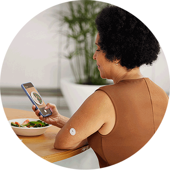 Person wearing a continuous glucose monitor on the upper arm sits at a table with a meal, using a smartphone to view food-related content.
