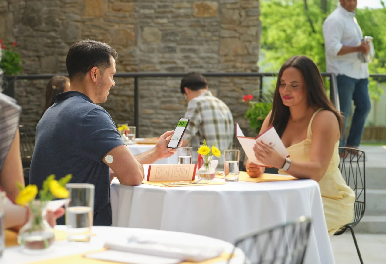 Hombre sentado con una mujer afuera mirando lecturas de glucosa en su teléfono.
