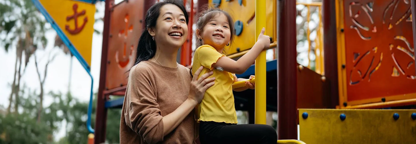 Một người đang giúp trẻ leo lên thiết bị sân chơi có các tấm nhiều màu sắc ở ngoài trời.