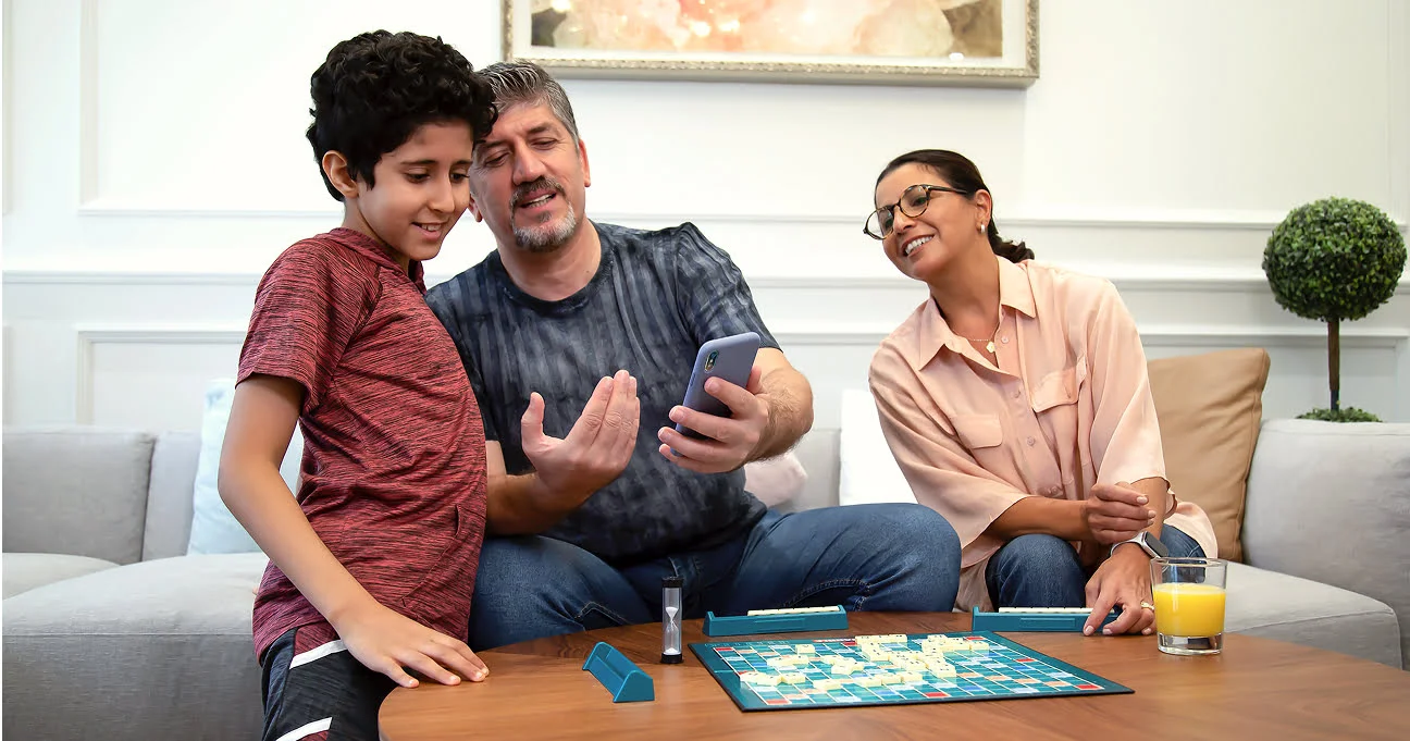 A father shows his son his smartphone while playing a board game with the family in their living room.