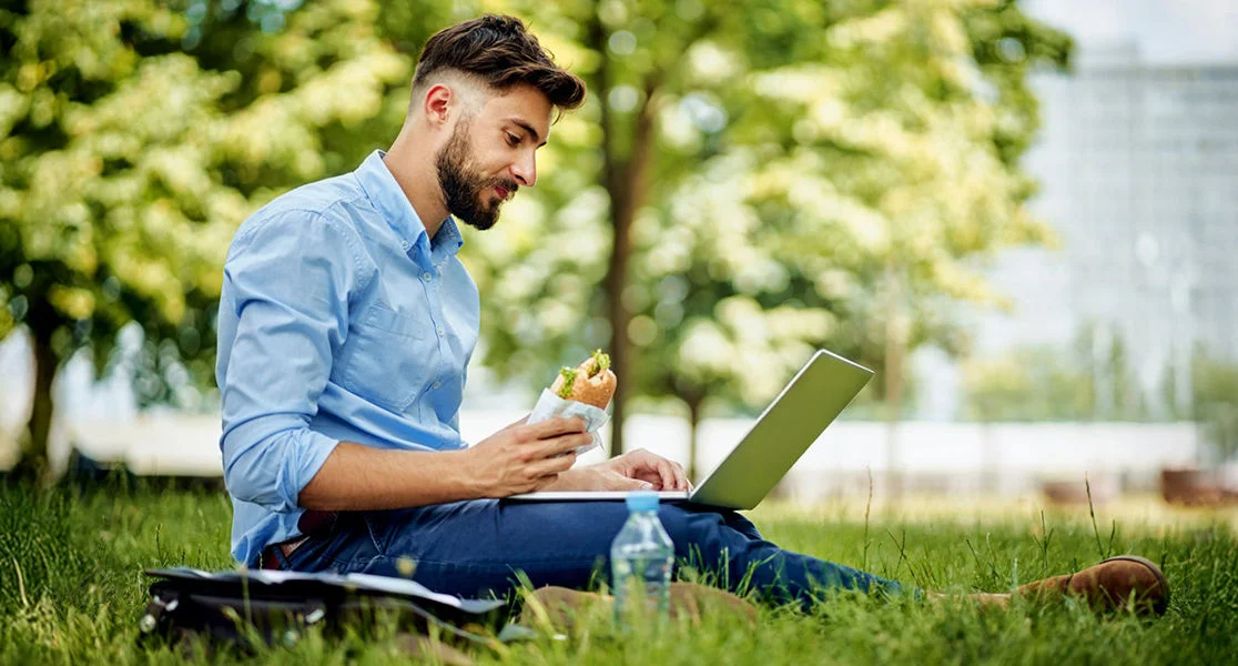 Person sitting on grass using a laptop while eating a sandwich outdoors in a park setting.