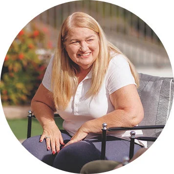 Person with long light-colored hair seated on a patio chair outdoors near landscaping and a fence.