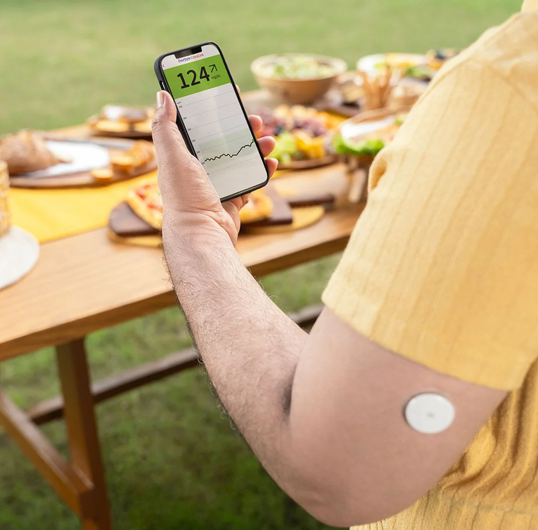 A person sitting outside at a picnic looking at their smartphone with a glucose sensor on their arm.