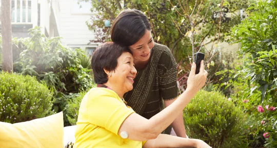 一名長者坐在椅子上，右手臂後側佩戴 FreeStyle Libre2 傳感器，與身旁一名年輕女士頭靠頭微笑看著 LibreLink 應用程式上的葡萄糖數值，背景為綠意盎然的庭院。
