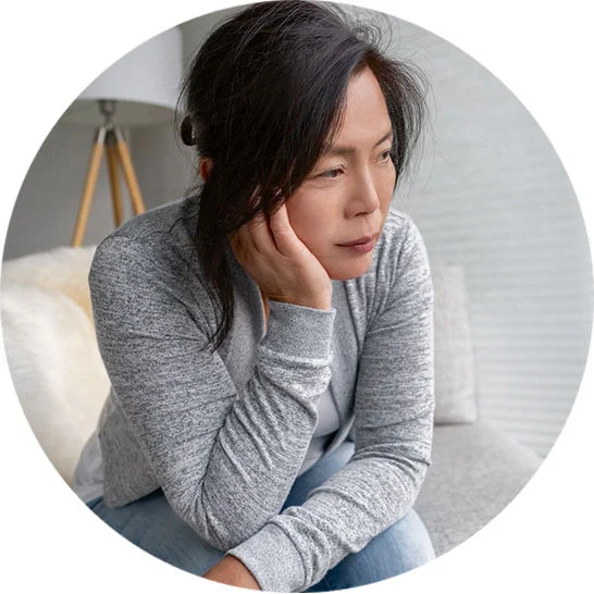 Person in a gray sweater sitting on a couch, resting head on hand and looking to the side in a bright room.