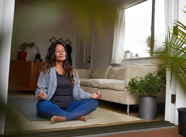 Mujer practicando meditación en casa, sentada con las piernas cruzadas y los ojos cerrados, reflejando calma y autocuidado