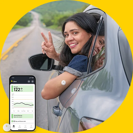 Person leaning out of a car window on an open road displays two wearable glucose sensors and makes a peace sign, with a glucose monitoring app shown on a smartphone screen.