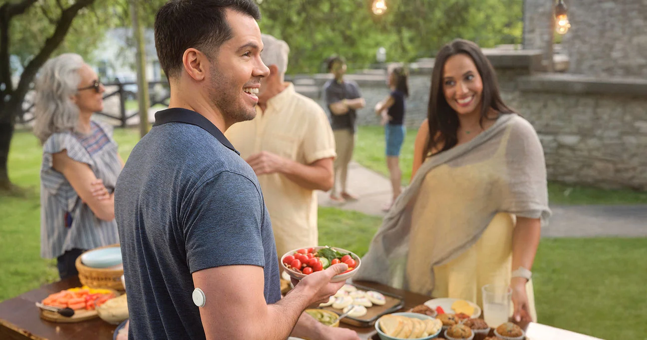A man wearing a FreeStyle Libre sensor chooses healthy vegetables from a buffet at an outdoor social gathering.