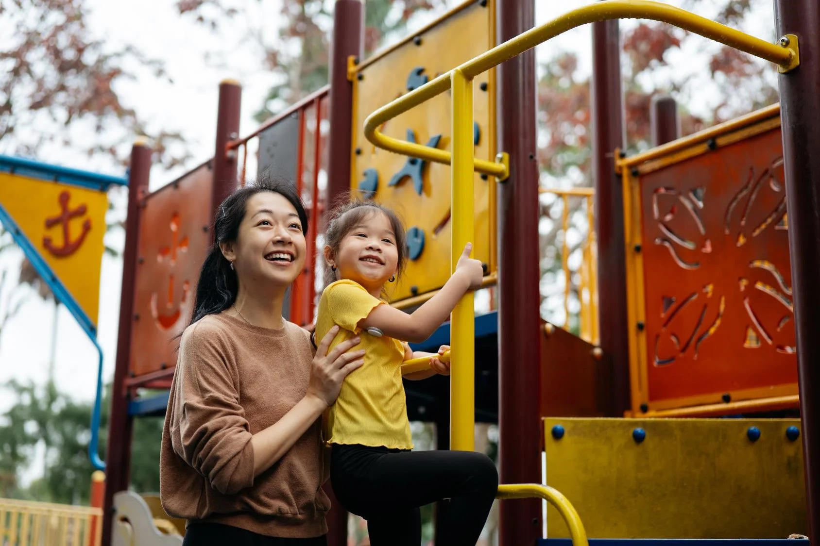 Adult holding a young child on a playground climbing structure with bright panels and bars in the background.