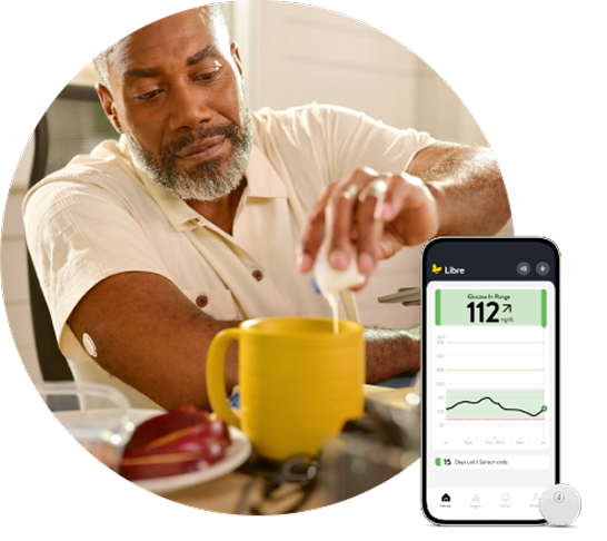 Person wearing a continuous glucose monitor on the upper arm pours creamer into a yellow mug while the FreeStyle Libre app displaying glucose data is shown on a smartphone nearby.
