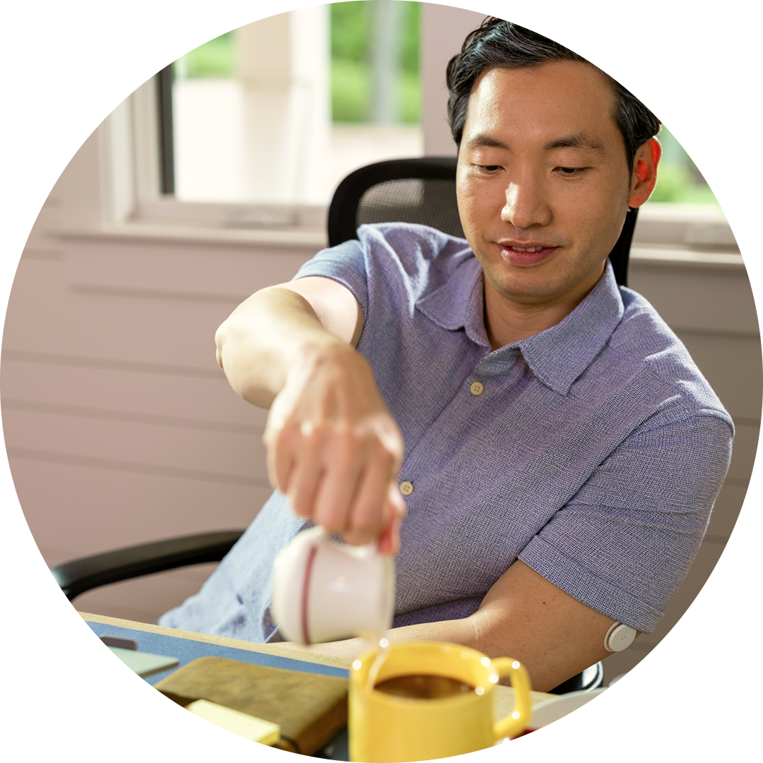 một người ngồi tại bàn trong nhà đang rót chất lỏng vào ly, cạnh màn hình hiển thị thiết bị đọc FreeStyle Libre 2