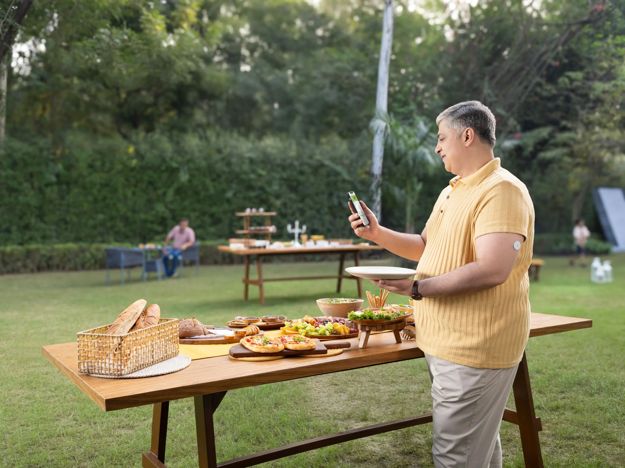 Man wearing a FreeStyle Libre sensor on the back of his arm checks his glucose reading on a smartphone while socializing with friends at an outdoor gathering.