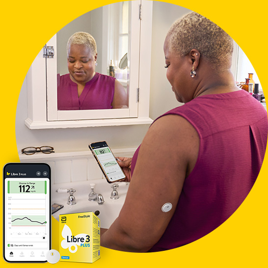 A woman checks glucose readings on a smartphone while wearing a sensor on the upper arm near a bathroom sink.
