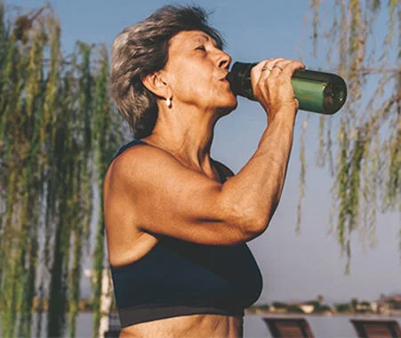 Person in athletic wear drinking water from a reusable bottle outdoors near trees and a waterfront.