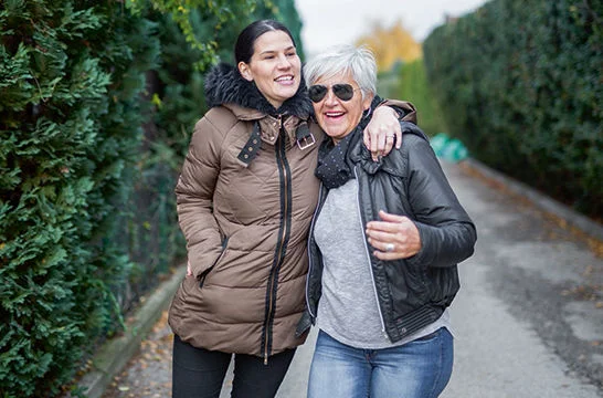 Two people walking arm in arm along a tree-lined pathway bordered by tall hedges.