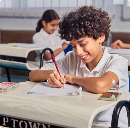 A kid sitting in his school desk with a smartphone clipped to the handlebars with LibreLinkUp opened.