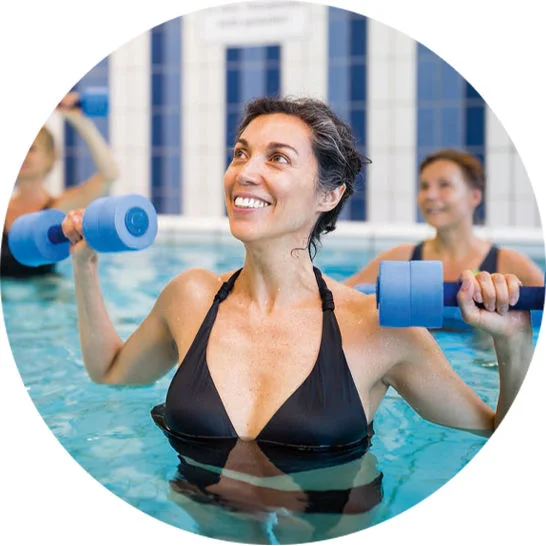 Person participating in a water aerobics class, holding foam dumbbells in a swimming pool while others exercise nearby.