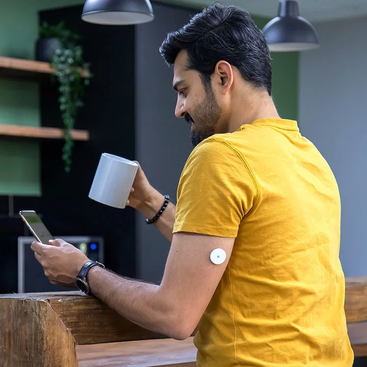 A man checks his phone while holding a mug at a counter, with a FreeStyle Libre sensor on his upper arm.