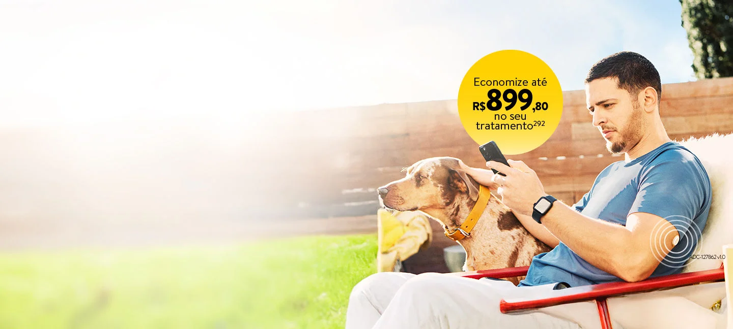 Homem sentado ao ar livre usando o smartphone e vestindo o sensor FreeStyle Libre no braço enquanto interage com um cachorro