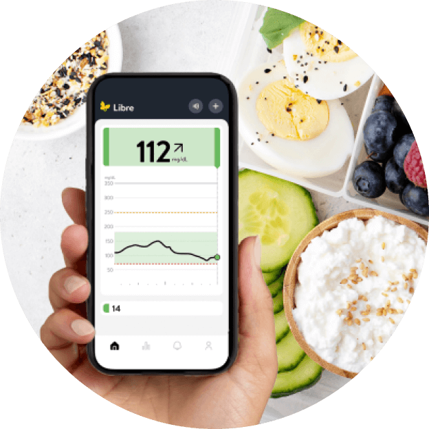 Persona sosteniendo un teléfono con la app FreeStyle Libre abierta, mostrando un nivel de glucosa estable junto a alimentos saludables como frutas y yogur.