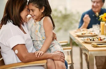mãe segurando a filha no colo em uma mesa de refeição com o seu sensor FreeStyle Libre no braço