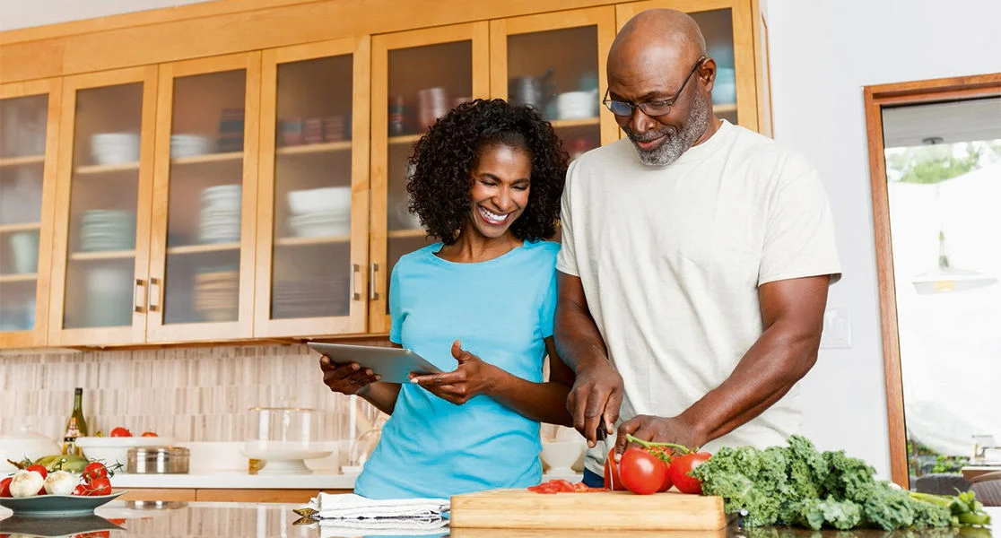 A couple preparing vegetables on a kitchen countertop while following a recipe on a tablet.