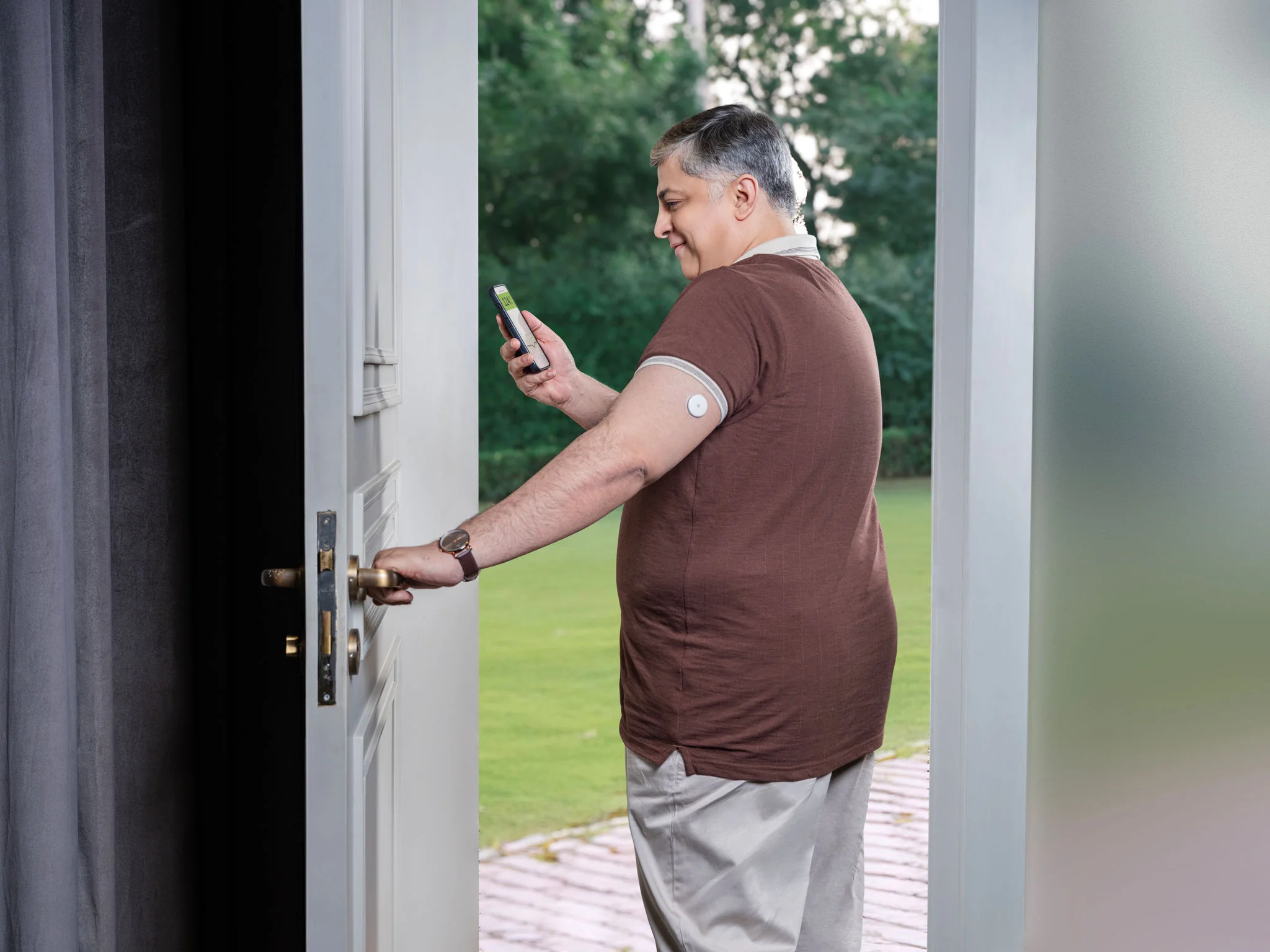 Man wearing a FreeStyle Libre sensor on his upper arm checks his glucose reading on a smartphone while sitting at a home office desk with a laptop and lunch.