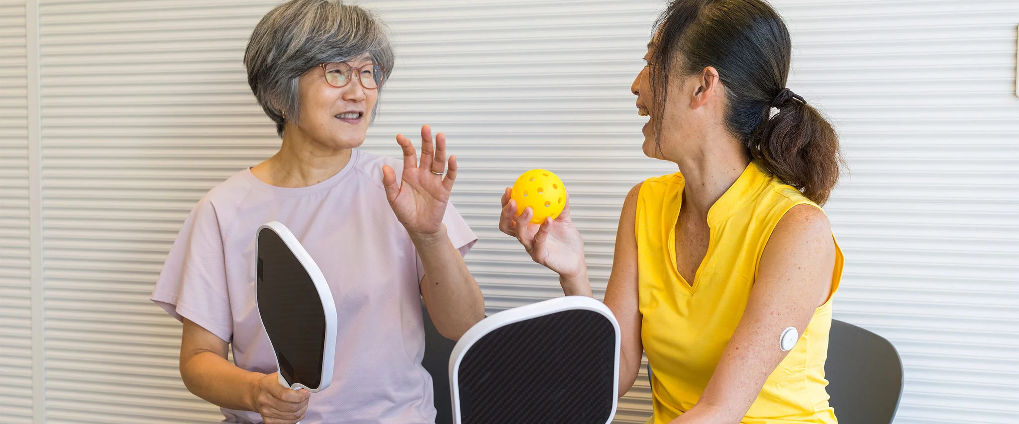 Two woman playing pickleball indoors, with one wearing a FreeStyle Libre sensor and holding a yellow perforated ball.