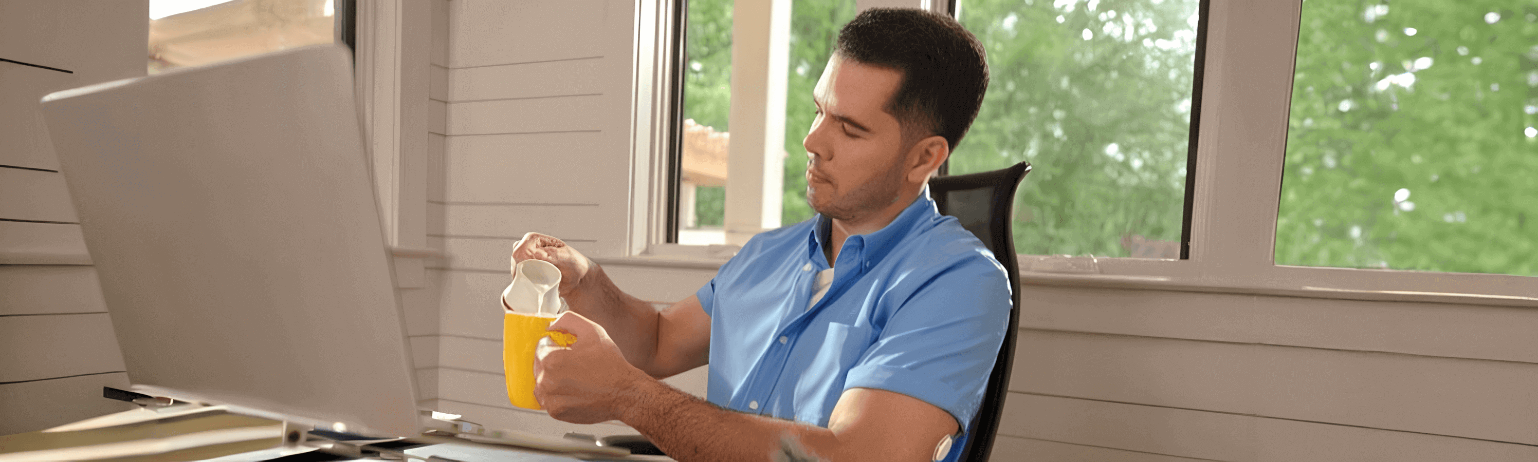 Homem sentado à mesa de trabalho despeja leite em uma caneca; ao fundo há janelas com vegetação e ele usa o sensor FreeStyle Libre 2 Plus no braço.
