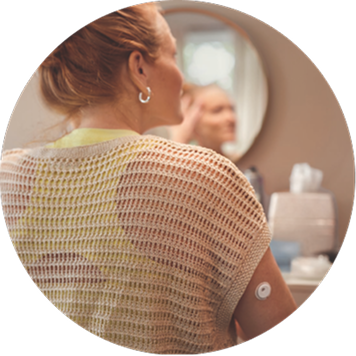 A woman seated indoors with a continuous glucose monitor sensor visible on the upper arm near a vanity mirror.