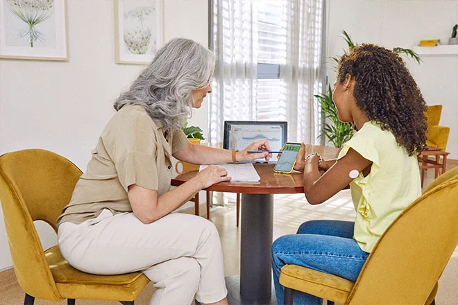 Senhora ensinando uma jovem sobre o aplicativo FreeStyle LibreLink. Ao fundo notasse uma tela com a pataforma Libre View