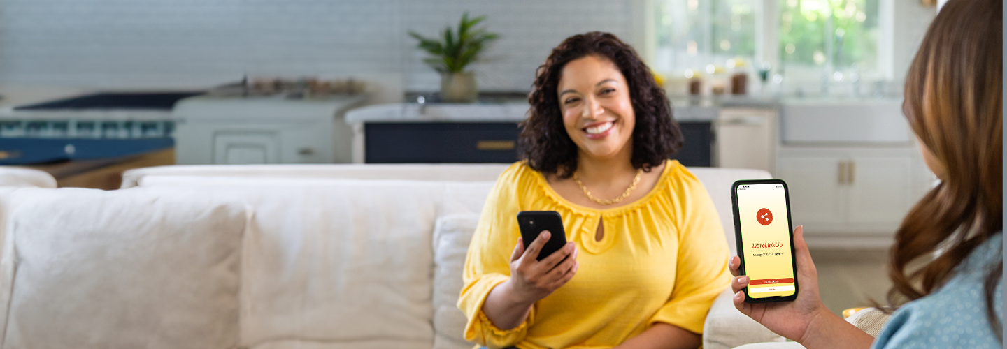 Person at home holding a smartphone while another phone displays the LibreLinkUp app, illustrating sharing glucose data with caregivers.