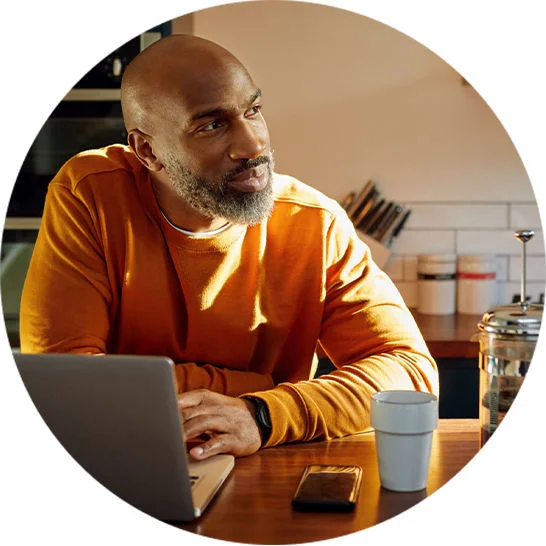 Person seated at a kitchen table using a laptop, with a smartphone, mug, and coffee press nearby.