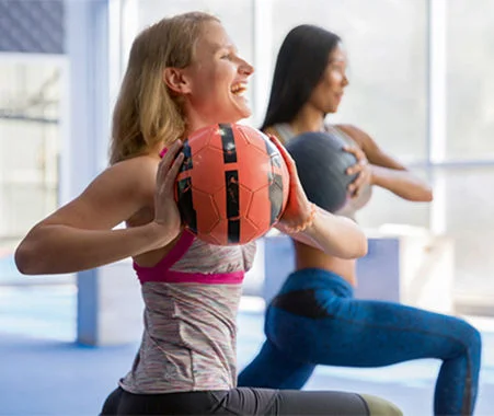 Two people performing medicine ball exercises in a bright fitness studio with large windows.