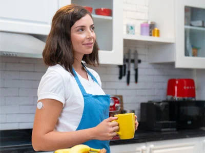 A happy woman wearing a FreeStyle Libre sensor, standing in the kitchen.
