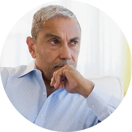 Person seated indoors with hand resting near chin in a thoughtful pose against a bright background.