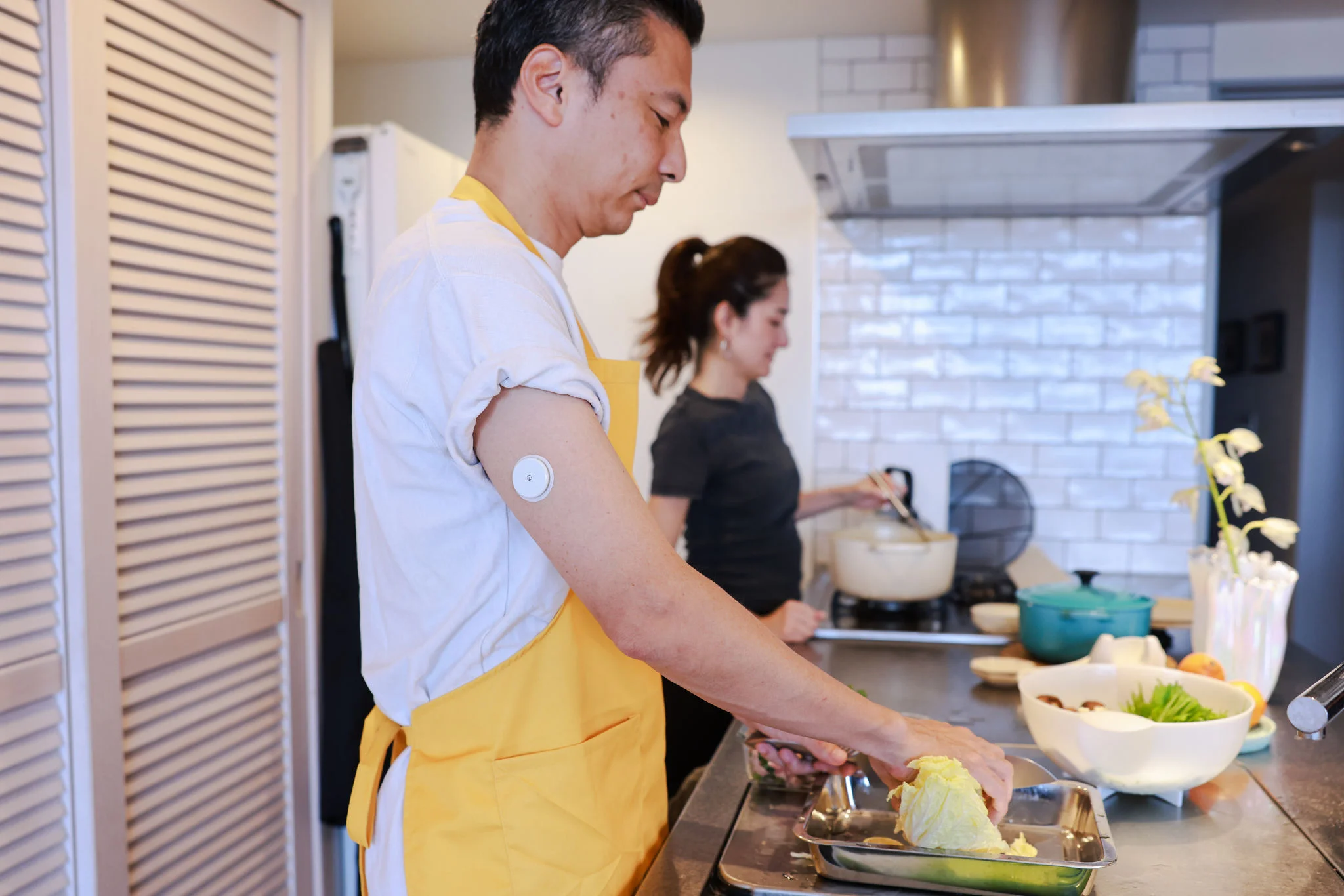 Person wearing a glucose monitor preparing food in a kitchen while another person cooks in the background.