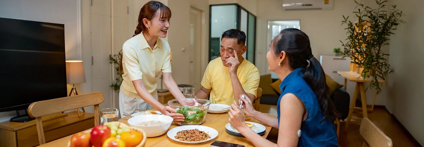 Três pessoas compartilham uma refeição saudável com salada, frutas e pratos equilibrados à mesa.