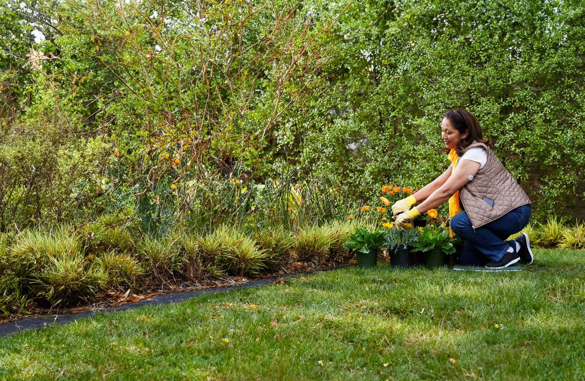 Mulher ajoelhada em um jardim cuidando de plantas com um sensor FreeStyle Libre no braço.