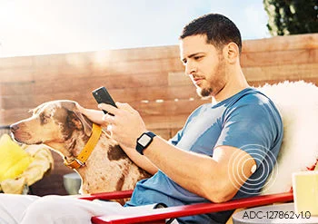 Homem sentado ao ar livre usando o celular enquanto acaricia um cachorro, com o sensor FreeStyle Libre visível no braço.