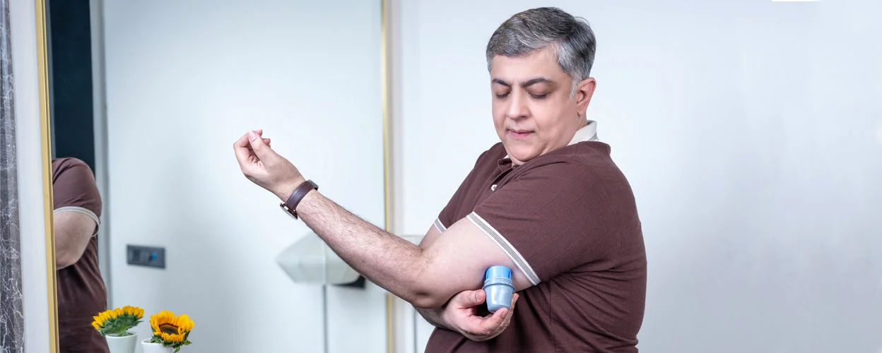 A man applying his FreeStyle Libre sensor on  the upper back of his left arm. near a window in a bright room..