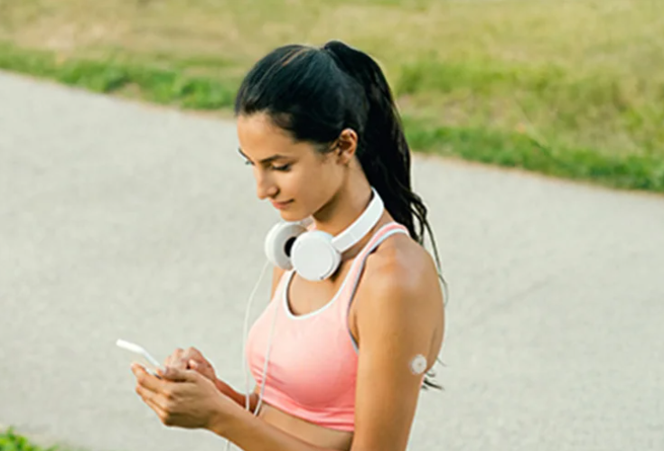 Mulher com fones de ouvidos apoiados no pescoço, verificando seu smartphone após realizar uma atividade física. Ela usa um sensor FreeStyle Libre em seu braço