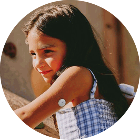 Child in a blue-and-white checkered sleeveless top outdoors, with a small round medical sensor attached to the upper arm.